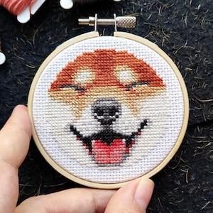 May include: A cross-stitch embroidery hoop featuring a smiling dog face. The dog's fur is a mix of brown and tan, with a black nose and a red tongue sticking out. The embroidery is set in a wooden hoop.