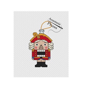 May include: A cross-stitch pattern featuring a Nutcracker soldier with a red and gold uniform, a white hat, and a brown beard. The pattern is titled "Anastasia Pruess".