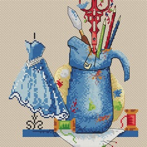 May include: A blue ceramic pitcher filled with art supplies, including scissors, pencils, and a paint scraper. A blue dress form with a blue dress is to the left of the pitcher. A spool of red thread and a piece of white fabric with pins are in the foreground.