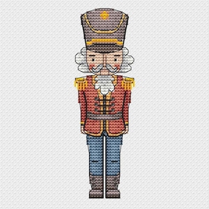 Puede incluir: Diseño de punto de cruz de un soldado cascanueces. El cascanueces lleva una chaqueta roja con charreteras doradas, pantalones azules y un sombrero alto. El diseño tiene una estética vintage y artesanal, para decoración navideña.