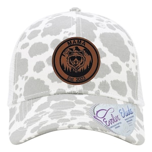 May include: A white and gray trucker hat with a brown leather patch that says "Mama" with a bear wearing sunglasses inside a mountain scene. The patch also says "Est. 2023".