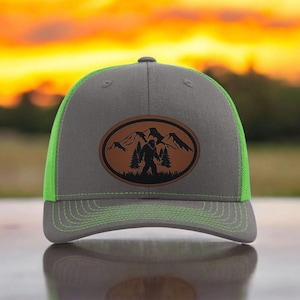 May include: A gray and green trucker hat with a brown leather patch featuring a silhouette of Bigfoot standing in front of mountains.