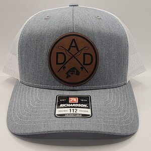 May include: A gray and white trucker hat with a brown leather patch that says "A.D.D." with a fishing rod and fish design. The hat is adjustable and has the Richardson logo on the back.