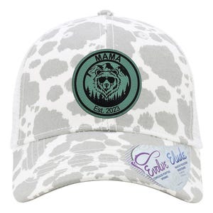May include: A white and gray trucker hat with a black and green patch that says "Mama Est. 2023" and features a bear with a mountain scene. The hat has a cow print pattern.