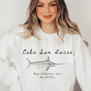 K&ouml;nnte beinhalten: Wei&szlig;er Pullover mit einem schwarzen grafischen Aufdruck eines Schwertfisches und dem Text "Cabo San Lucas, Baja California Sur -Rey Del Mar-."