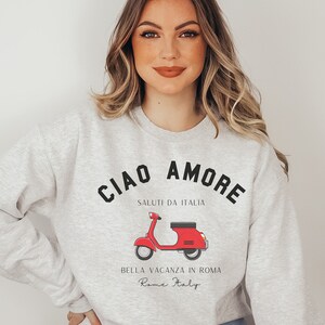 Op de afbeelding: Grijs sweatshirt met een zwarte grafische print van een rode scooter en de tekst "Ciao Amore", "Saluti da Italia", "Bella Vacanza in Roma" en "Rome Italy".