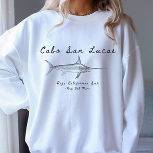 K&ouml;nnte beinhalten: Wei&szlig;er Pullover mit einem schwarzen grafischen Aufdruck eines Schwertfisches und dem Text "Cabo San Lucas, Baja California Sur -Rey Del Mar-."