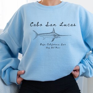 K&ouml;nnte beinhalten: Hellblauer Pullover mit einer schwarzen Grafik eines Schwertfisches und dem Text "Cabo San Lucas, Baja California Sur -Rey Del Mar-."