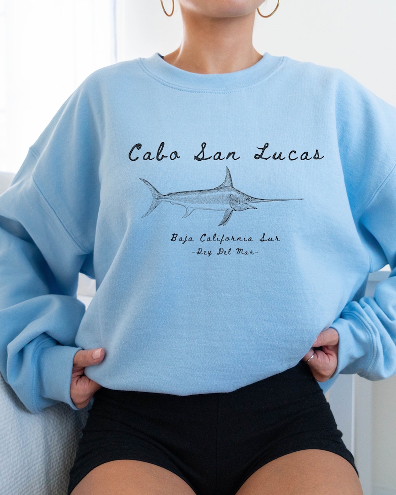 K&ouml;nnte beinhalten: Hellblauer Pullover mit einer schwarzen Grafik eines Schwertfisches und dem Text "Cabo San Lucas, Baja California Sur -Rey Del Mar-."