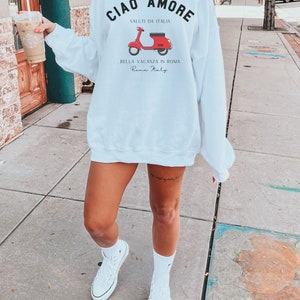Op de afbeelding: Wit sweatshirt met een rode scooter afbeelding en de tekst "Ciao Amore Saluti da Italia Bella Vacanza in Roma Rome Italy".