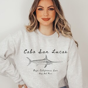 K&ouml;nnte beinhalten: Ein graues Sweatshirt mit einer Grafik eines Schwertfisches und dem Text "Cabo San Lucas, Baja California Sur - Rey Del Mar -" auf der Vorderseite.