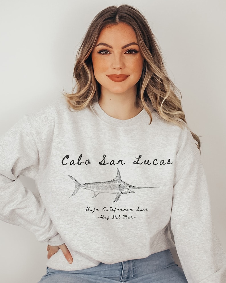 K&ouml;nnte beinhalten: Ein graues Sweatshirt mit einer Grafik eines Schwertfisches und dem Text "Cabo San Lucas, Baja California Sur - Rey Del Mar -" auf der Vorderseite.