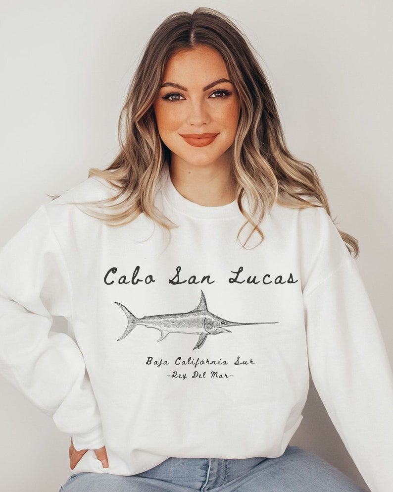 K&ouml;nnte beinhalten: Wei&szlig;er Pullover mit einem schwarzen grafischen Aufdruck eines Schwertfisches und dem Text "Cabo San Lucas, Baja California Sur -Rey Del Mar-."