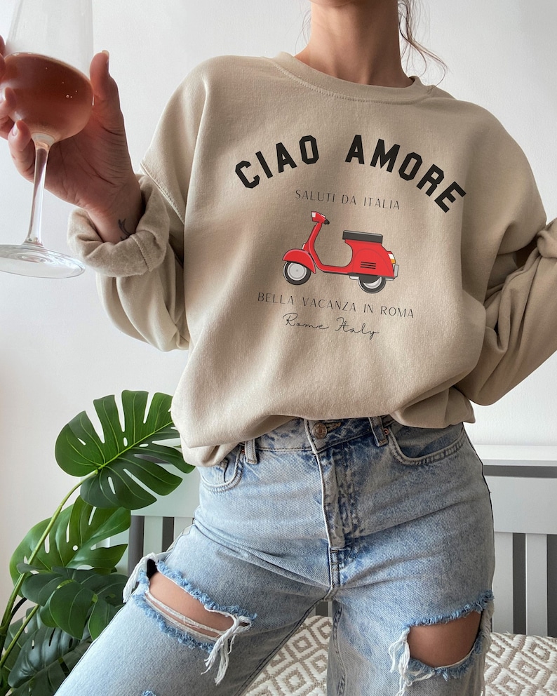 Op de afbeelding: Een beige sweatshirt met de tekst "Ciao Amore" en een rode scooter afbeelding. De tekst onder de scooter luidt "Saluti da Italia" en "Bella Vacanza in Roma Rome Italy".