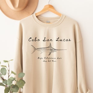 K&ouml;nnte beinhalten: Ein beiger Pullover mit einer schwarzen Grafik eines Schwertfisches und dem Text "Cabo San Lucas, Baja California Sur - Rey Del Mar - "
