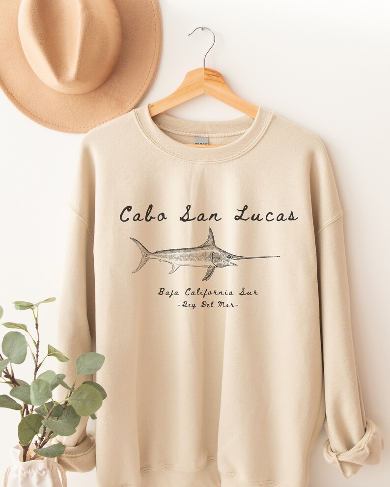 K&ouml;nnte beinhalten: Ein beiger Pullover mit einer schwarzen Grafik eines Schwertfisches und dem Text "Cabo San Lucas, Baja California Sur - Rey Del Mar - "