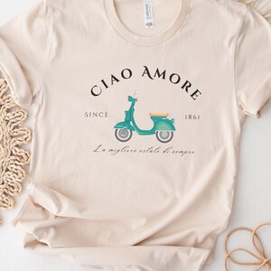 Op de afbeelding: Een beige T-shirt met een afbeelding van een turquoise scooter en de tekst "Ciao Amore Since 1861 La migliore estate di sempre".