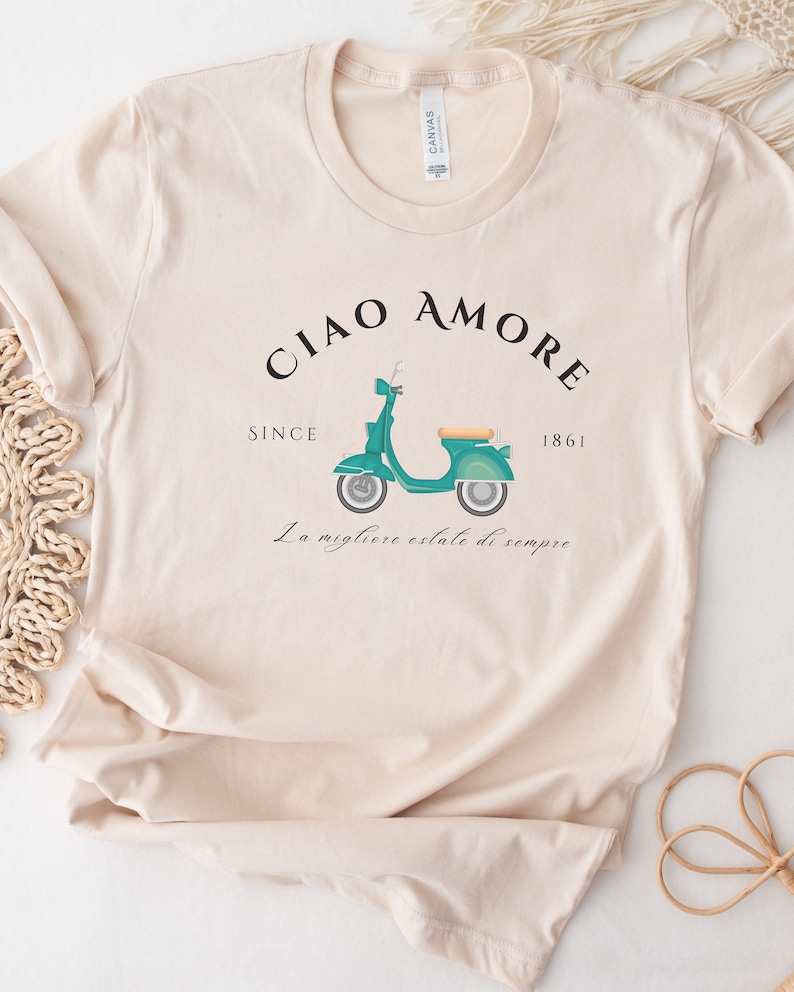 Op de afbeelding: Een beige T-shirt met een afbeelding van een turquoise scooter en de tekst "Ciao Amore Since 1861 La migliore estate di sempre".