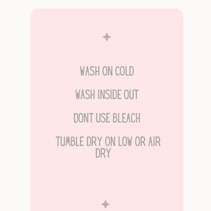 Może przedstawiać: R&oacute;żowa karta z tekstem "SHIRT CARE" u g&oacute;ry i listą instrukcji pielęgnacyjnych: "WASH ON COLD", "WASH INSIDE OUT", "DONT USE BLEACH", "TUMBLE DRY ON LOW OR AIR DRY". Na dole karty znajduje się tekst "magical wishing Co."