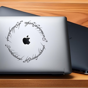 Op de afbeelding: Een zilveren laptop met een zwart Apple-logo en een zwarte vinyl sticker met de inscriptie van de Ene Ring uit De Heer der Ringen.