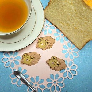 Puede incluir: Tres pines de esmalte de color dorado con ranas de dibujos animados con la lengua fuera. Las ranas son marrones y tienen ojos grandes y redondos. Los pines están sobre un mantel de encaje rosa con fondo azul.