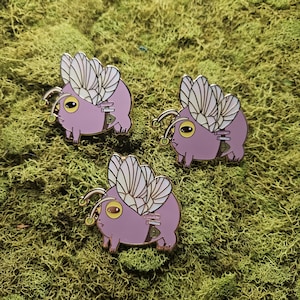 Puede incluir: Tres pines de esmalte que representan una criatura redonda y linda con cuerpos de color púrpura claro, grandes alas de color crema y detalles dorados. Cada pin tiene una pequeña bolsa de dinero y una antena dorada. Los pines están sobre musgo verde.