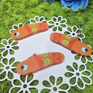May include: Three orange keyrings with a fish design, featuring green circles and a blue eye. The keyrings are on a white, floral-shaped paper doily, set against a green moss background.