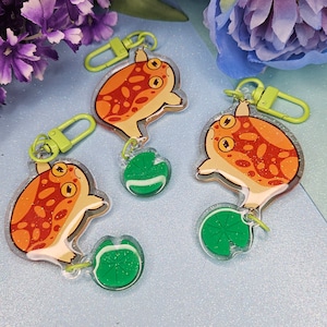 May include: Three acrylic frog keyrings with orange and brown patterns, yellow eyes, and green clasps. Each frog is attached to a green lily pad charm. The keyrings are on a light blue surface.
