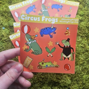 May include: Square stickers with the text "Circus Frogs" and illustrations of frogs in circus attire. The frogs are green and blue, with various accessories like party hats, bow ties, and juggling pins. The stickers are on an orange background.