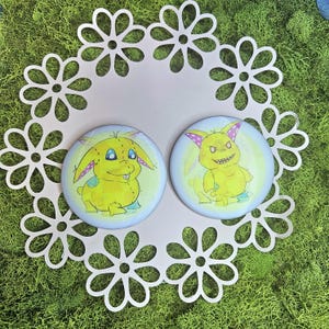 May include: Two round, yellow cartoon monster pins with blue and pink accents. The pins are on a light pink, daisy-shaped doily, set against a background of green moss.