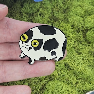 May include: A black and white enamel pin of a cartoon frog with large yellow eyes. The frog has a black and white spotted pattern, similar to a cow.