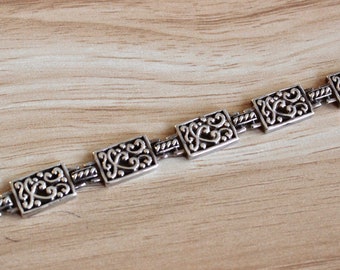 May include: A silver bracelet with a chain link design and rectangular charms featuring intricate floral patterns.