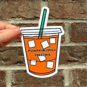 May include: A sticker of an orange drink with ice cubes and a green straw. The text "PUMPKIN SPICE SPEECHIE" is printed on the front of the drink. The sticker is held in front of a brick wall.