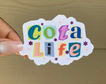 May include: A white sticker with the words "cocoa life" in colorful, cut-out letters. The letters are in different colors, including pink, blue, green, yellow, and purple. There are small purple flowers and white stars scattered around the letters.
