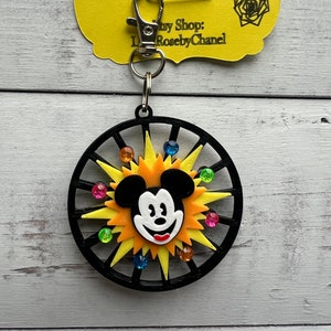 Może przedstawiać: Czarno-żółty brelok z postacią Myszki Miki z wzorem gwiazdy i kolorowymi kryształkami. Brelok ma srebrny metalowy pierścień i srebrny metalowy zatrzask. Nazwa sklepu Etsy "Lena & Rose" jest nadrukowana na żółtej metce przymocowanej do breloka.