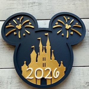 Peut inclure: Décoration en forme de tête de Mickey Mouse, bleu marine et dorée, avec un château et des feux d'artifice. Le chiffre "2026" est affiché. Il s'agit probablement d'un article commémoratif ou d'une décoration de fête.