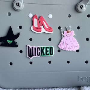May include: A gray tote bag with a variety of colorful patches, including a black witch with green skin, a pair of red shoes, a pink dress with a crown, and a black and green "WICKED" patch.