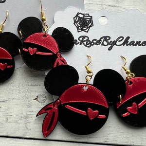 May include: A pair of black and red Mickey Mouse-shaped earrings with a red bandana and a heart design. The earrings are on a white card with the text "Rose By Chanel".