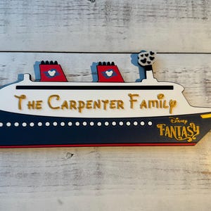 May include: A wooden ship-shaped sign with the text "The Carpenter Family" in gold lettering. The ship is white with a navy blue hull and red accents. The words "Disney Fantasy" are also visible.