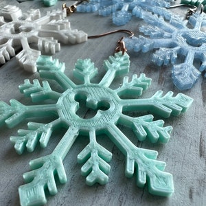 May include: A pair of snowflake earrings with a light blue, white, and teal color scheme. The snowflakes are detailed and have a 3D appearance.