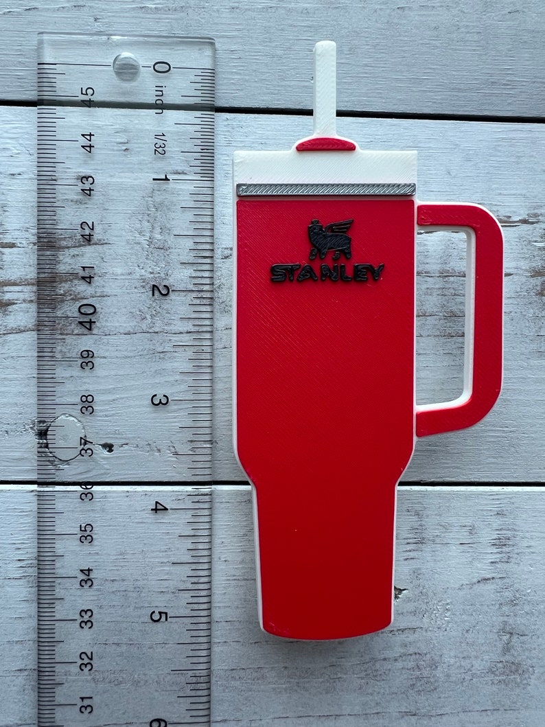 Close up of red Stanley tumbler charm shown next to a US ruler.  It is approximately 5 inches from tip of straw to bottom of cup.  It is about 2.25 inches wide at the handle.