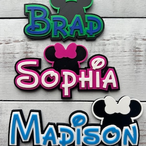 May include: Three personalized Mickey Mouse and Minnie Mouse themed name plaques. The top plaque is green and blue with the name "Brad". The middle plaque is pink and white with the name "Sophia". The bottom plaque is blue and white with the name "Madison".