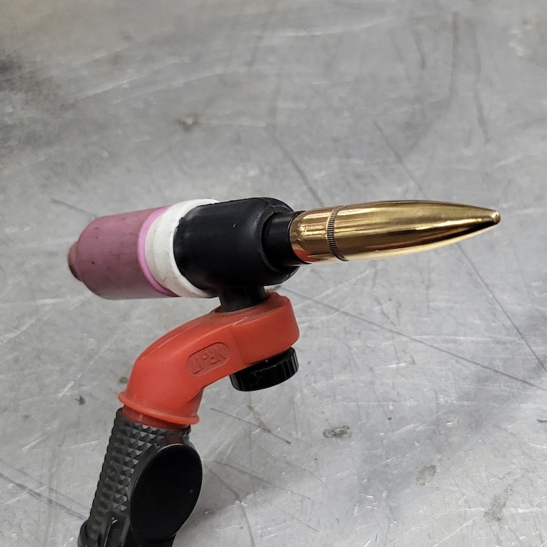 May include: A close-up of a welding torch with a gold-coloured tip. The torch is attached to a red and black handle with a black grip.