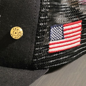 May include: Black trucker hat with a mesh back and an embroidered American flag on the right side. A gold-colored bullet casing with the text "S&B 45 AUTO" is attached to the front of the hat.