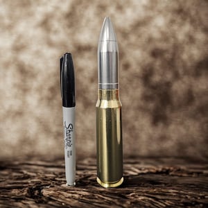 Op de afbeelding: Een zwarte Sharpie fine point marker naast een grote, metalen kogel. De kogel heeft een zilveren punt en een messingkleurige behuizing. De achtergrond is een getextureerd, bruin oppervlak.