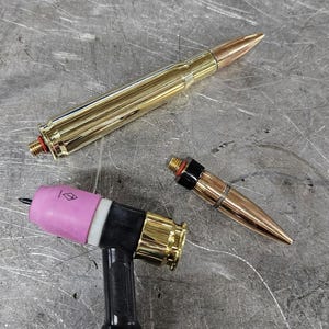 May include: A TIG welding torch head with a pink ceramic nozzle, a brass back cap, and two bullet-shaped accessories. One accessory is a long brass cartridge casing, and the other is a shorter copper-coloured bullet tip with a black rubber ring.