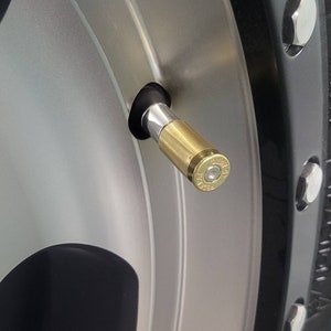 May include: A gold-colored tire valve stem cap shaped like a bullet casing. The cap is attached to a silver valve stem and is visible against a silver and black tire rim.