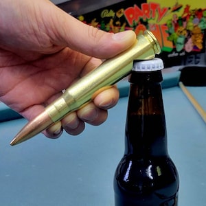 Può includere: Un apribottiglie dorato a forma di proiettile viene utilizzato per aprire una bottiglia di birra di vetro marrone.