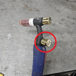May include: A TIG welding torch handle featuring a blue grip and a black neck. The torch is customised with two gold-coloured brass bullet casing caps, one at the top and one on the side, with the side cap highlighted by a red circle.