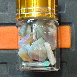 Könnte beinhalten: Ein kleines Glas mit einem goldfarbenen Deckel, gefüllt mit verschiedenen bunten Edelsteinen. Die Edelsteine sind hauptsächlich in Blau-, Grün- und Weißtönen gehalten. Das Glas befindet sich in einem dunkelgrauen Behälter mit einem orangefarbenen Akzent.